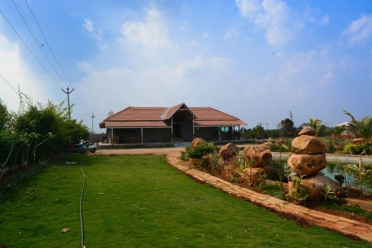 Single-storey tiled-roof farmhouse on a green lawn with a stone-edged path, boulders and pond under a blue sky near Hyderabad