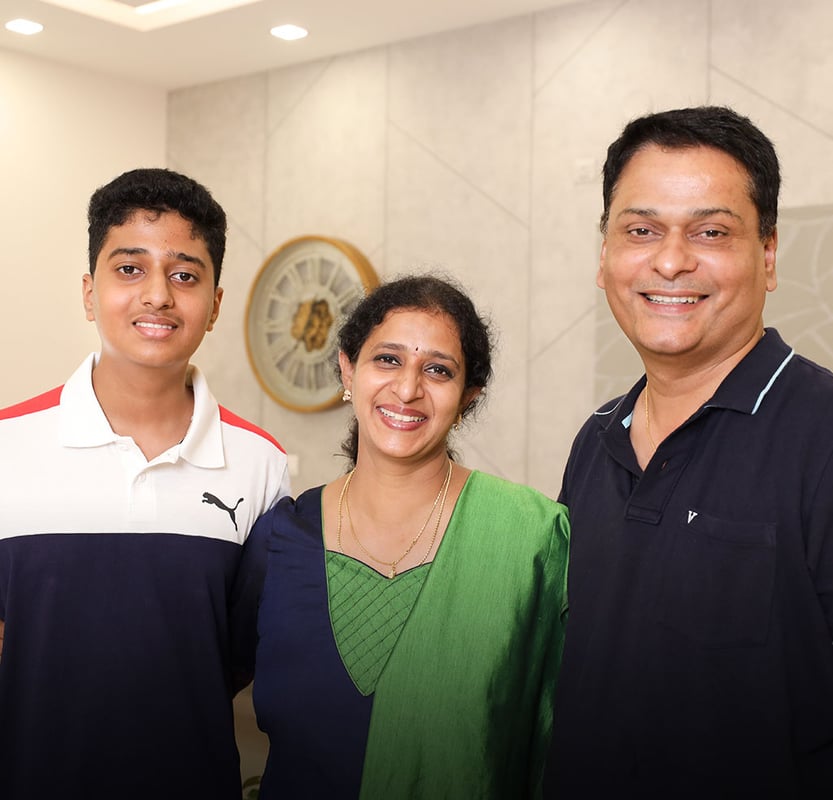 Rao family portrait of three smiling members in front of a beige wall with a round Roman-numeral wall clock