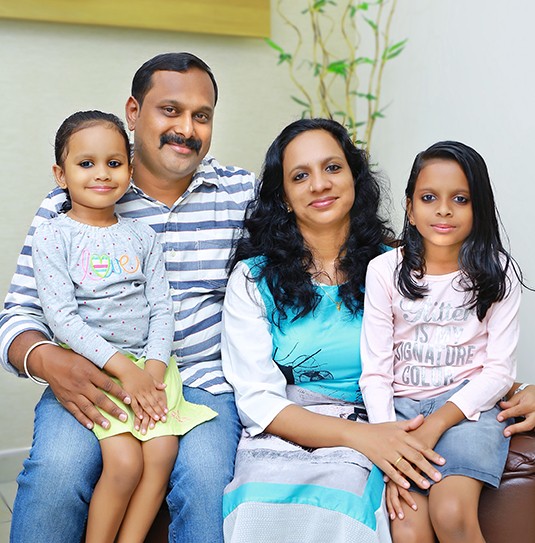 Appukkuttan family portrait of two parents and two daughters seated together against a soft neutral wall