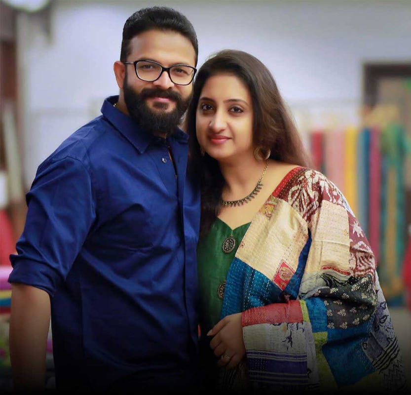 Portrait of a smiling couple, the man in navy shirt and woman in patchwork-sleeve outfit, against a soft library backdrop