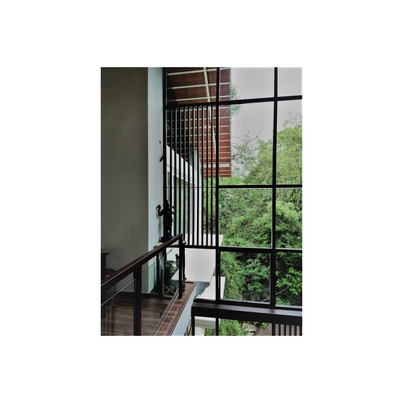 Double-height stairwell with tall black grid-framed windows and a dark metal railing framing leafy garden views, Hyderabad